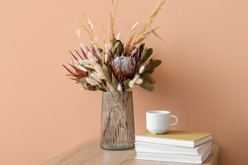 Vase with beautiful dried flowers, books and cup on table near beige wall