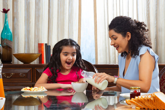 Mother pouring milk into bowl with chocos for her daughter during breakfast at home