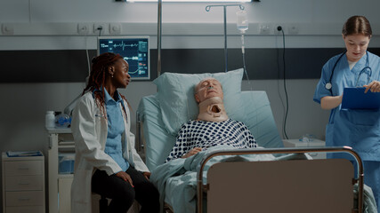 Multi ethnic medical staff checking on patient healthcare in hospital ward bed at intensive care. African american doctor helping old man with cervical neck collar and oxygen tube