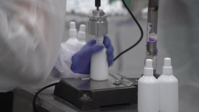 Close up of screwing bottle caps on white antiseptic plastic bottles without labels by semi-automatic machine and gloved hands with out of focus table and same bottles on background