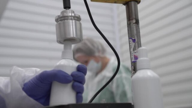 Close up of screwing two bottle caps by semi-automatic machine and gloved hands with out of focus worker and white walls on background. Bottles without labels