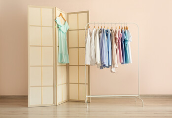 Stylish folding screen and rack with female clothes near wall in room