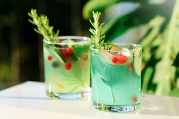 Two glasses with green lemonade made from lemon rosemary and mint in glasses on table at the terrace. Cold refreshing beverage for hot summer day. Green monstera leaves on background.