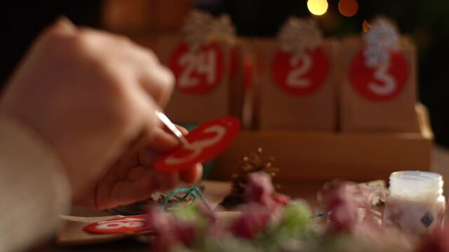 Close-up tracking shot of unrecognizable man writing number on red bag by brush and white paint on blurred background. Male making paper bags from kraft paper for advent calendar on Christmas Eve.