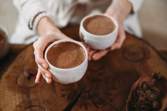 Hot handmade ceremonial cacao in white cups. Woman hands giving craft cocoa, top view on wooden table. Organic healthy chocolate drink prepared from beans, foam Giving cup on ceremony cozy atmosphere