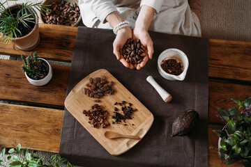 Woman hands holding organic cacao beans on wooden table, cocoa nibs, artisanal chocolate making in rustic boho style for ceremony. Degustation, Chocolate making with pounder close-up top view - obrazy, fototapety, plakaty