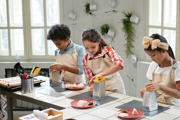 Group of children in aprons rubbing cheese on the plates while standing at the table, they cooking...