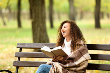Happy African-American woman reading book on bench in autumn park