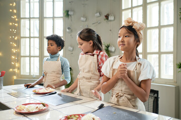 Group of children in aprons standing near the table with cutting board, they learning to cook at...
