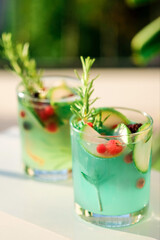 Two glasses with green lemonade made from lemon rosemary and mint in glasses on table at the terrace. Cold refreshing beverage for hot summer day. Green monstera leaves on background.
