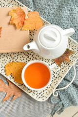 Tray with tea pot, cup of hot beverage and autumn leaves on sofa in room