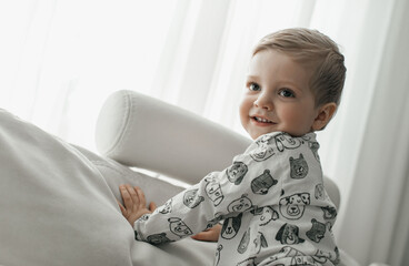Fototapeta premium A happy little child in a white jacket and brown pants lies comfortably on a white soft sofa and laughs against the background of a large bright window. 