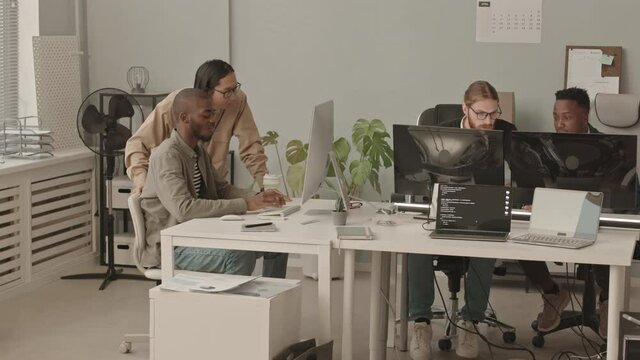 Panning shot of four young male programmers of diverse ethnicities working in team at contemporary office, typing on keyboards and talking in front of computer monitors