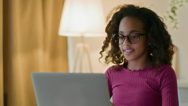 E-learning concept. Cheerful teen african american girl wearing eyeglasses typing on laptop, studying distantly at home