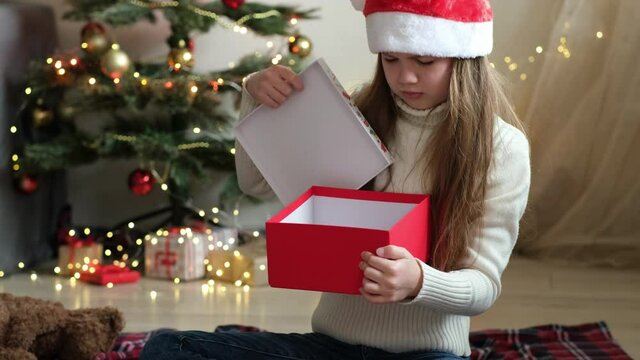 Little girl in Santa hat opens a Christmas gift and gets upset. Received the wrong present she dreamed of. Child on background of decorated tree disappointed and feeling unhappy with holiday gift