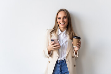 Portrait of a satisfied young businesswoman using mobile phone while holding cup of coffee to go over white background - obrazy, fototapety, plakaty