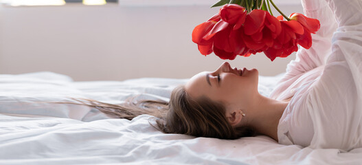 a young beautiful Caucasian woman lies on a bed in a white shirt holds red tulips in her hands and...