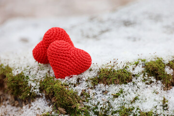 Two knitted red hearts on the snow and green moss in a forest. Valentine's card, symbols of love, background for romantic event in winter, Christmas celebration