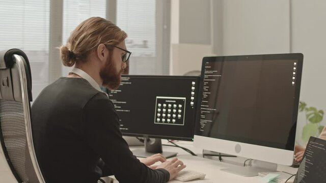 Handheld waist up slowmo shot of serious programmer with beard typing program code on computer sitting at modern open space office with colleagues