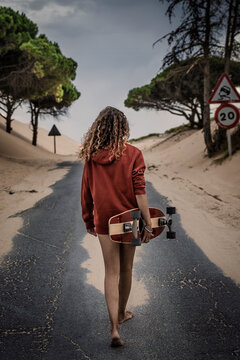 Chica de espaldas con sudadera roja caminando con su patinete en la carretera de las dunas de Valdevaqueros, Tarifa, Cádiz, Andalucia, España