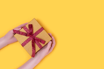 Hands hold gift box tied with red glittering ribbon on a yellow background. Festive composition with copy space.