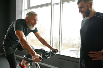Fototapeta na wymiar Elderly man on bike with young trainer in gym