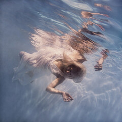 A woman with blond hair in a white dress swims underwater as if flying in zero gravity