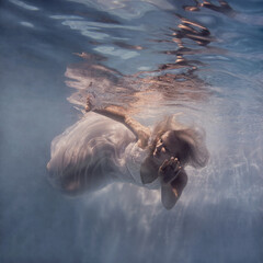 A woman with blond hair in a white dress swims underwater as if flying in zero gravity