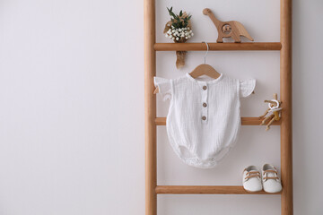 Fototapeta na wymiar Baby bodysuit, shoes, toys and small bouquet on ladder near white wall. Space for text