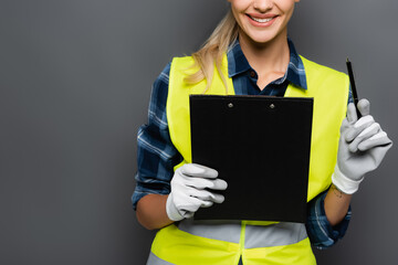 Cropped view of cheerful blonde builder in gloves and safety vest holding pen and clipboard isolated on grey