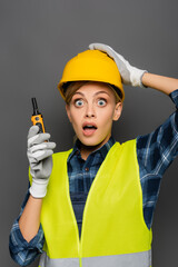 Scared builder in hardhat and safety vest holding walkie talkie and looking at camera isolated on grey