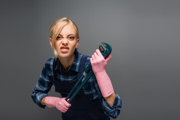 Angry plumber in overalls holding pipe wrench and looking at camera isolated on grey