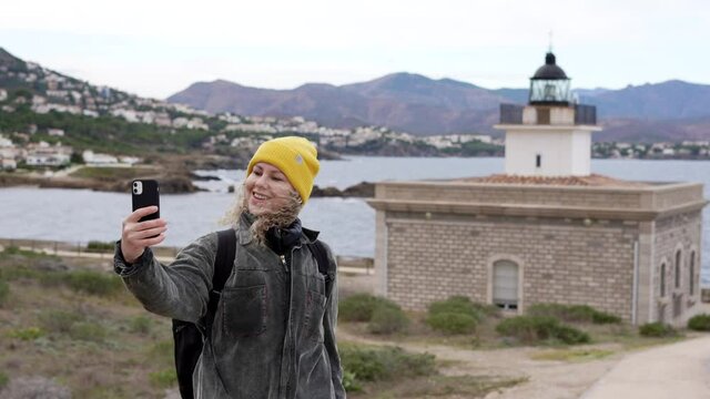 Woman With Yellow Hat Making A Video Call On The Windy Beach To Show Her Friends The Views Of The Sea And The Lighthouse.