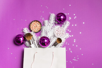 Gift paper bag with fir tree branches, balls, shining decorations and decorative cosmetic products on trendy purple background top view. Christmas cosmetic sale and promo banner.