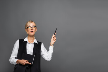 Pensive businesswoman holding notebook and pointing with hand isolated on grey