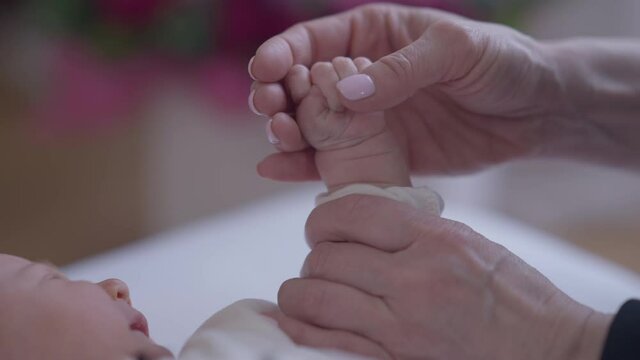 Close-up female Caucasian hand caressing fist of newborn crying girl indoors. Unrecognizable loving mother stroking little fingers as displeased little child lying in bed