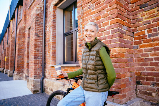 Trendy Laughing Middle Age Blond Woman Walking With Bicycle, With Reusable Coffee Cup In Hand. Acrive Female Going To Work Place By Her Bike In Morning.