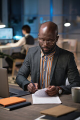 African businessman in eyeglasses sitting at his workplace and signing documents while working at...