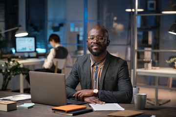 Fototapeta na wymiar Portrait of African businessman in eyeglasses smiling at camera while working at his workplace with laptop till late evening at office