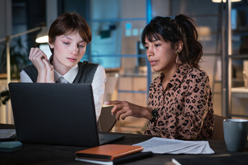 Businesswoman pointing at the screen of laptop and discussing online presentation with her colleague, they working at office till late evening