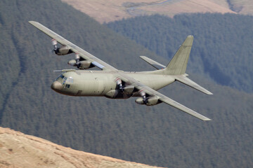 Green military C-130 Hercules flying low level with trees and hills behind. Low flying in Wales,...