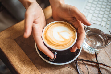 Closeup shot of an unrecognizable woman having a cup of coffee at a cafe. Lady's hands holding cup with sth heart-shaped. Female hands in warm sweater holding cup of coffee