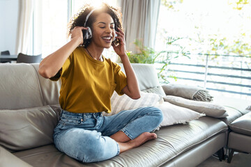 Motivated mature woman listening to music with headphones on her smartphone while dancing and singing on sofa at home.