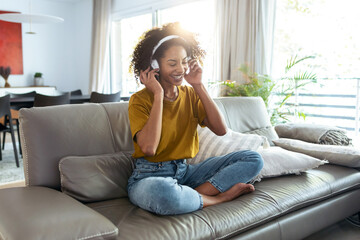 Fototapeta premium Motivated mature woman listening to music with headphones on her smartphone while dancing and singing on sofa at home.