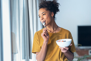 Beautiful mature woman eating cereals and fruits while standing next to the window at home. - Powered by Adobe