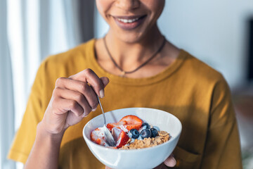 Beautiful mature woman eating cereals and fruits while standing next to the window at home.