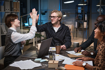 Young businessman giving a high-five to his colleague while they sitting at the table at meeting and working till late evening