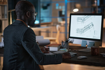 Rear view of African engineer sitting at the table in front of computer monitor and developing blueprint online during work at office