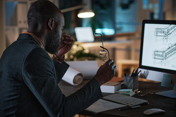 Tired African architect sitting at the table in front of the computer monitor with blueprint while working at office till night