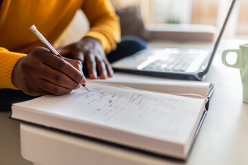 One mature man working at home and writing down some notes. Close up of an unrecognized black man at the flat sitting on sofa at home doing finances on his laptop, writing down some notes.